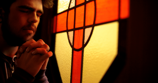 一个留着棕色短发、头戴头巾的人在一扇黄红相间的彩色玻璃窗旁合十祈祷.